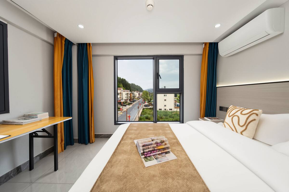 Una habitación de hotel con una cama, un escritorio, un ventanal con cortinas y un equipo de aire acondicionado.