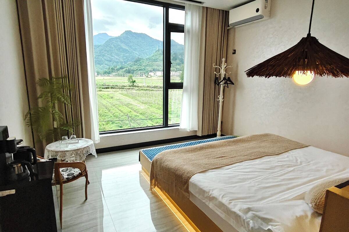 Una habitación de hotel moderna con un amplio ventanal que ofrece vistas a un paisaje montañoso. La habitación tiene una cama bien hecha, una mesita redonda pequeña con una silla y un escritorio con una silla.