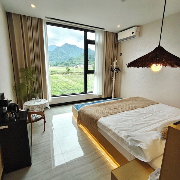 Una habitación de hotel moderna con un amplio ventanal que ofrece vistas a un paisaje montañoso. La habitación tiene una cama bien hecha, una mesita redonda pequeña con una silla y un escritorio con una silla.