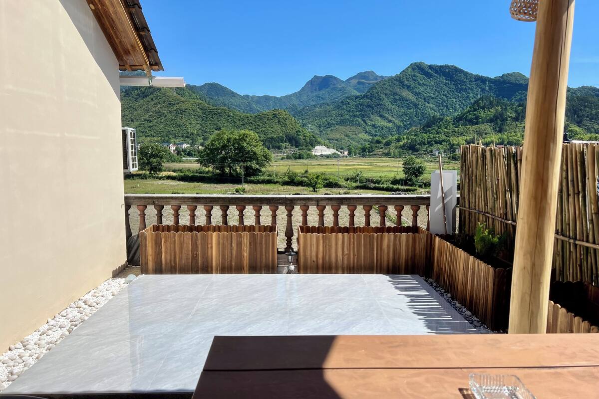 Un balcón con vistas a montañas, una valla de madera y una mesa con un recipiente de vidrio.