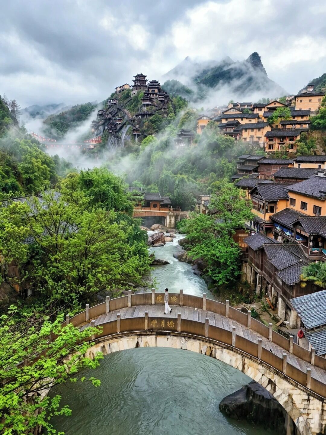 Un village traditionnel, avec une rivière, un pont et une montagne en arrière-plan.