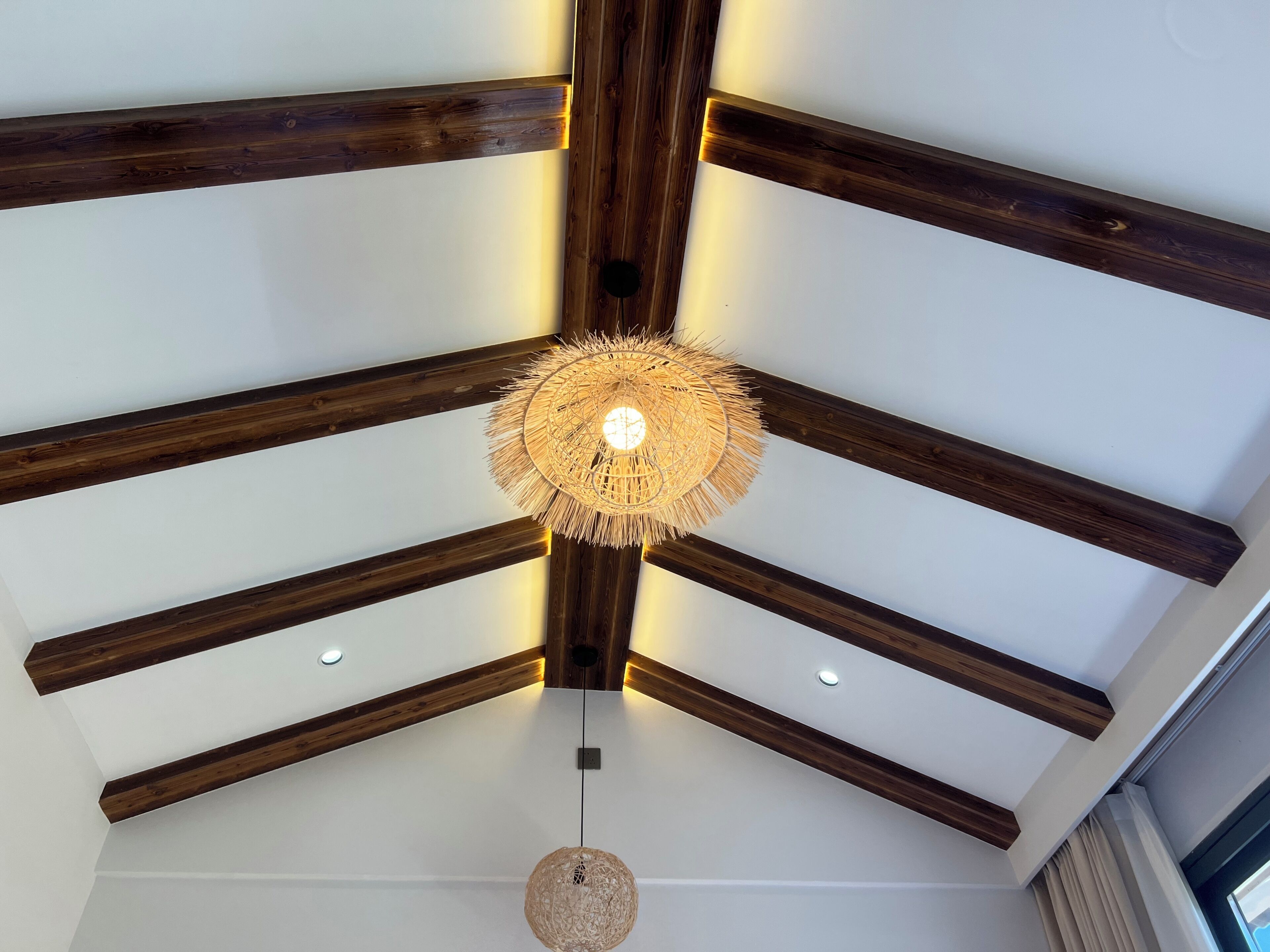 Una habitación con vigas de madera a la vista, un candelabro tejido en el techo, luces empotradas y una lámpara colgante.