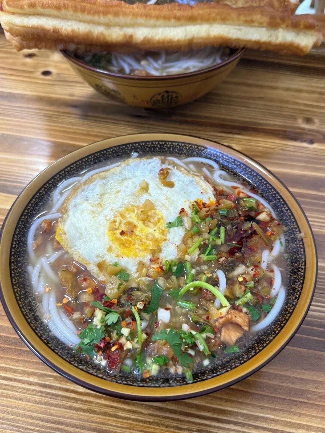 Un tazón de sopa de fideos con un huevo frito, decorado con hierbas y verduras, sobre una mesa de madera.