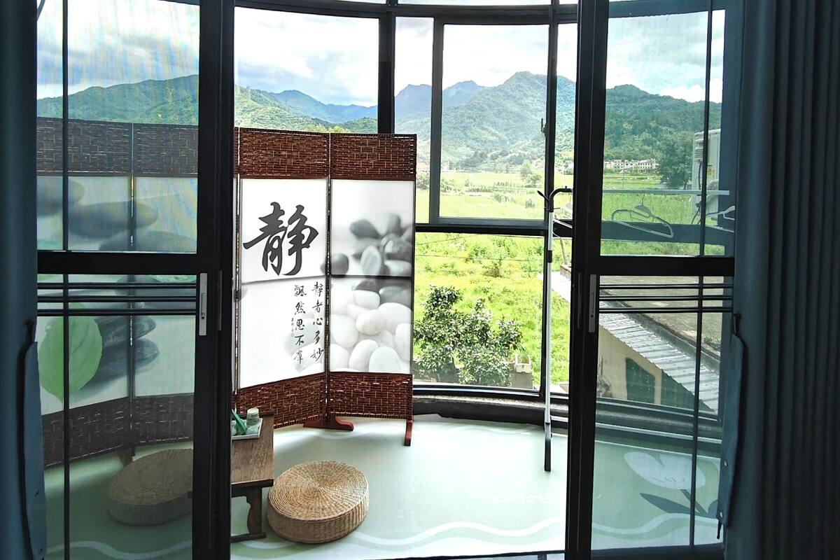 Habitación con amplios ventanales que ofrecen vistas a un paisaje verde y montañas. Se observa una pantalla japonesa tradicional con caligrafía y un taburete tejido.