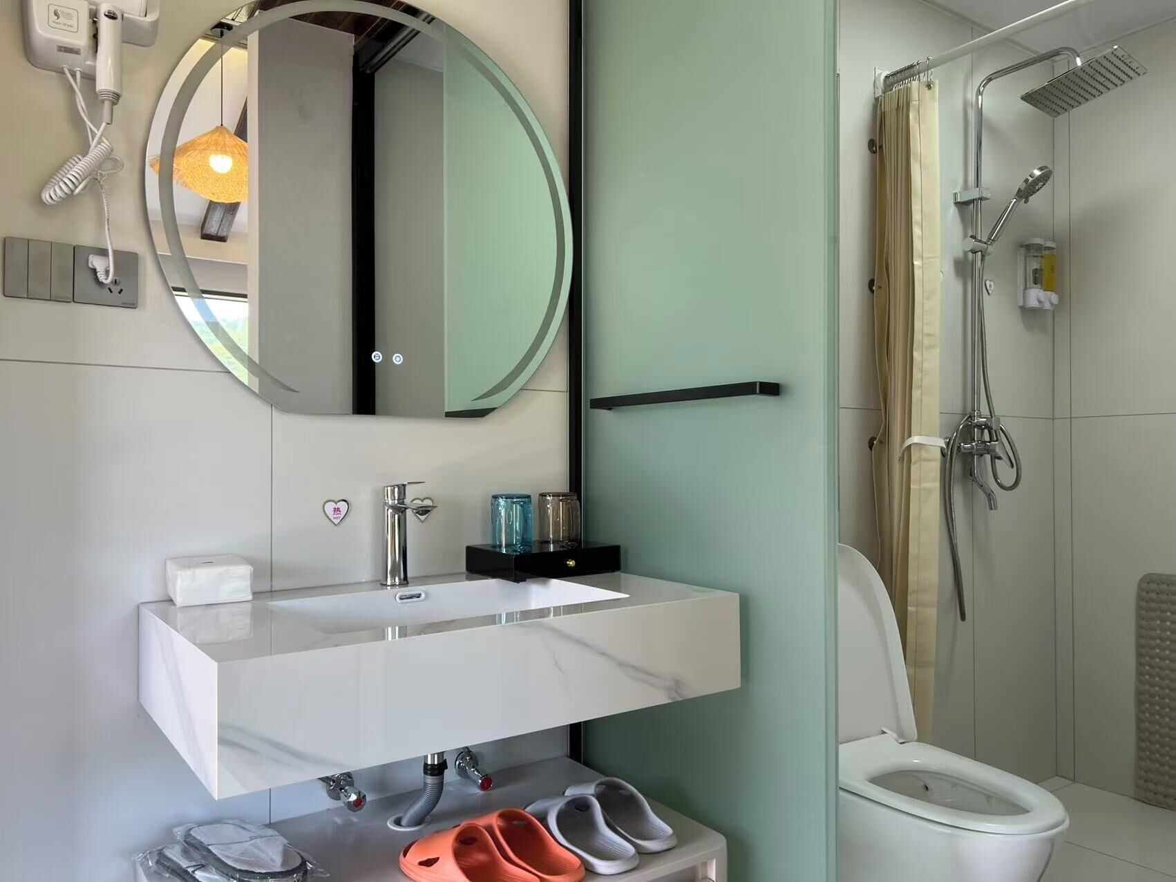 Ein modernes Badezimmer mit einem weißen Waschtisch, einem runden Spiegel, einer Duschkabine aus Glas und einer Toilette.