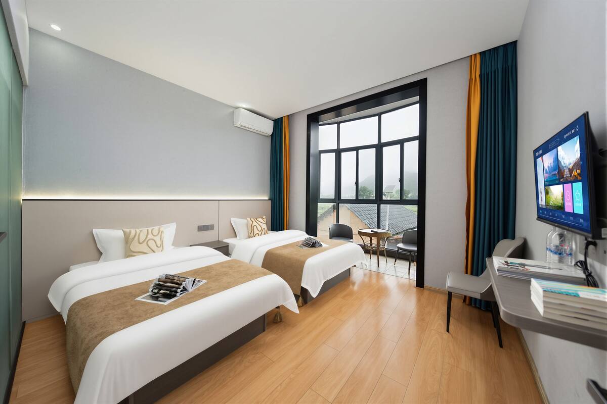 Habitación de hotel con dos camas, un escritorio, una silla, un televisor y un ventanal grande.