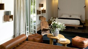 A modern hotel room with a large bed, a wooden chair, leather ottomans, a small table with a vase, and a view of greenery through the window. - My Rental Homes - Villa Susan with heated pool and large garden (Marrakesh)