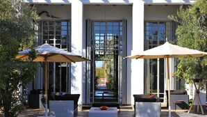 A modern outdoor seating area with two umbrellas, a white building with large windows, and a view of a water feature. - My Rental Homes - Villa Susan with heated pool and large garden (Marrakesh)