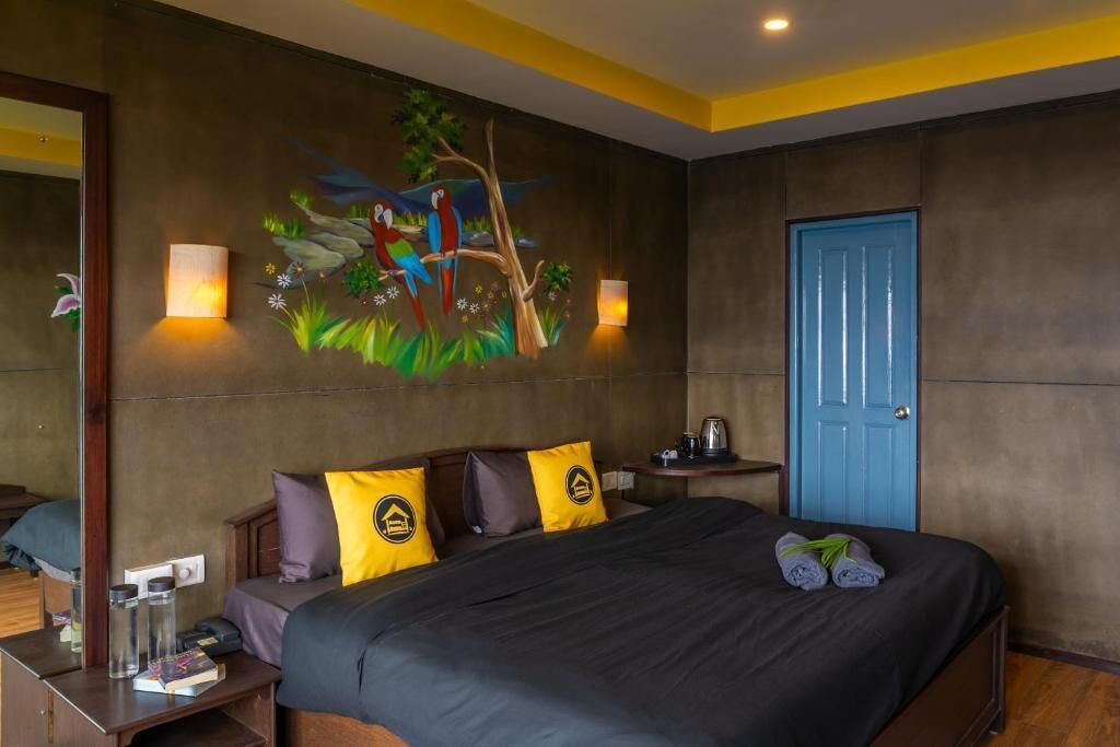 A hotel room with a bed, a nightstand, a lamp, a painting of parrots on the wall, and a blue door.