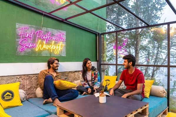 A group of people sitting on a blue sofa with yellow cushions, in a room with a green wall and a neon sign that reads "Find People on The Same Wave." - The Hosteller Munnar (Devikolam)