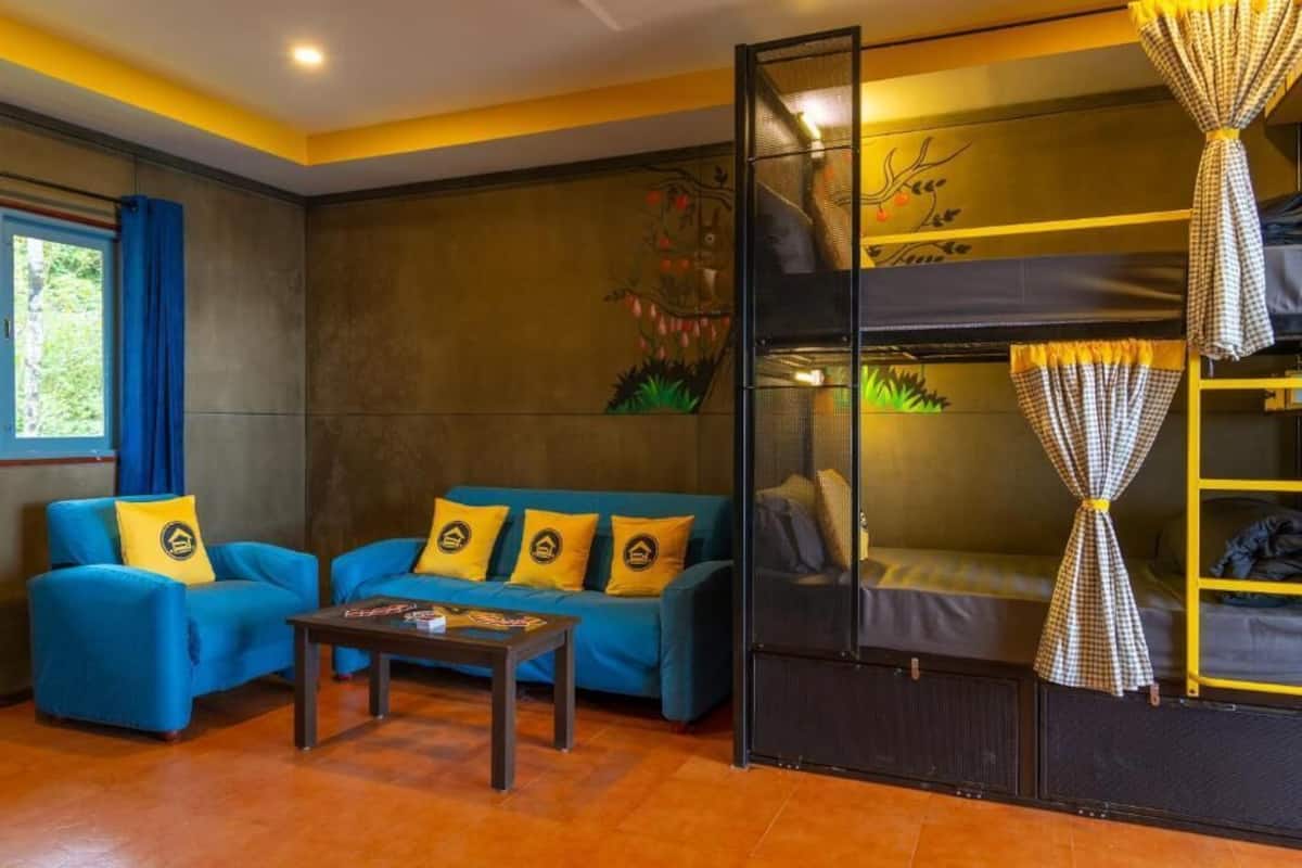 A room with a bunk bed, two blue sofas with yellow cushions, a small table, and a window with curtains.