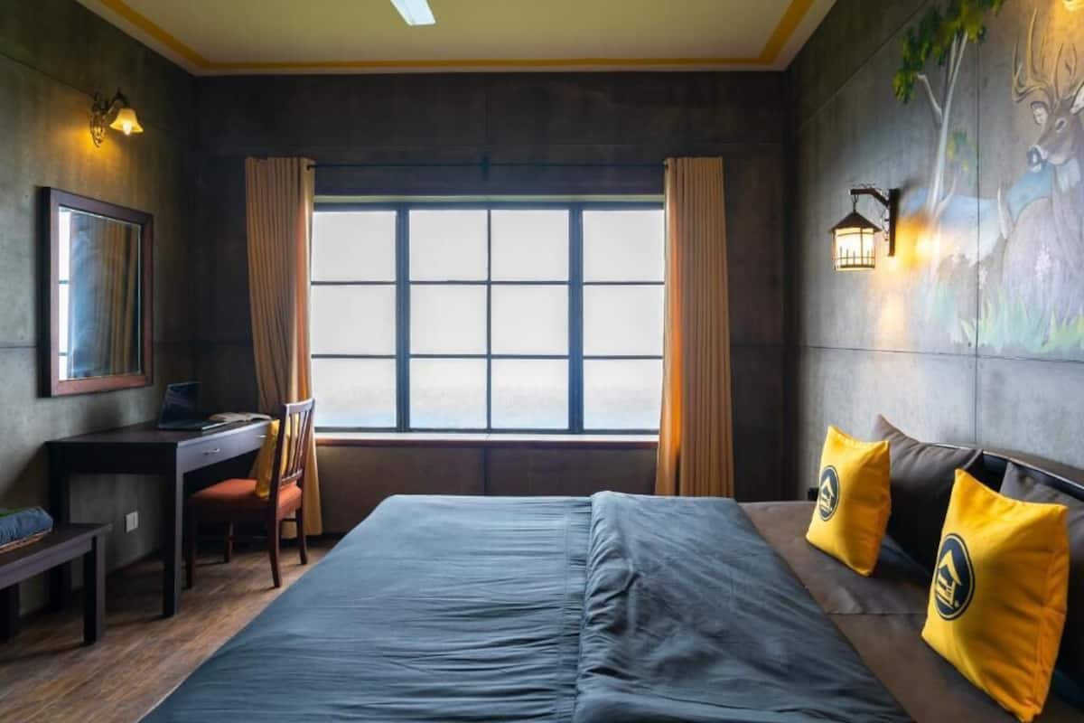A hotel room with a large bed, a desk, and a chair. There is a window with curtains, a wall-mounted lamp, and a painting of a deer.