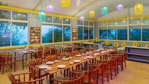 Een ruime eetruimte met houten tafels en stoelen, grote ramen en kleurrijke hanglampen.