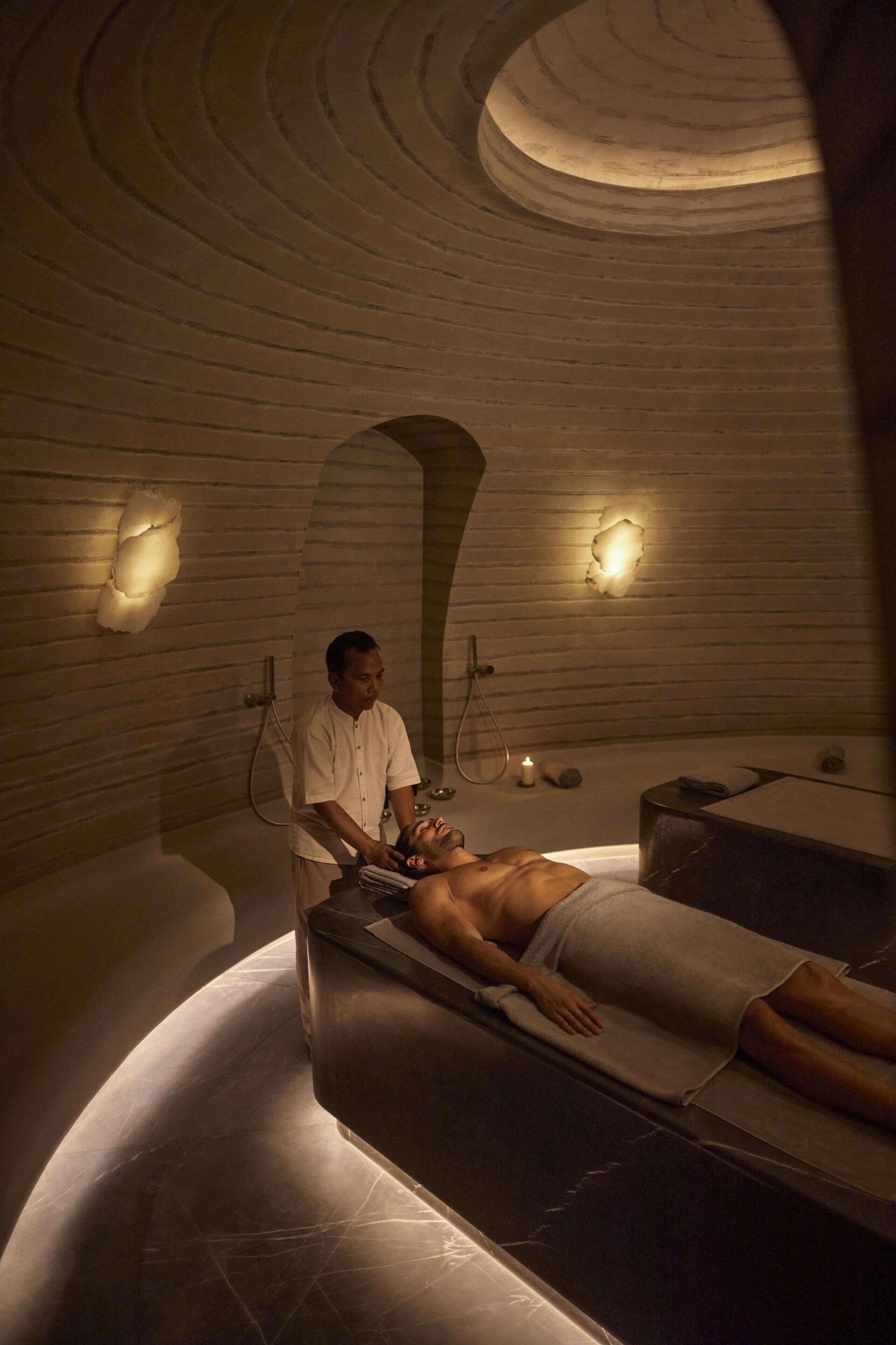 A spa room with a person lying on a massage table, a masseuse attending to them, and a bathtub.