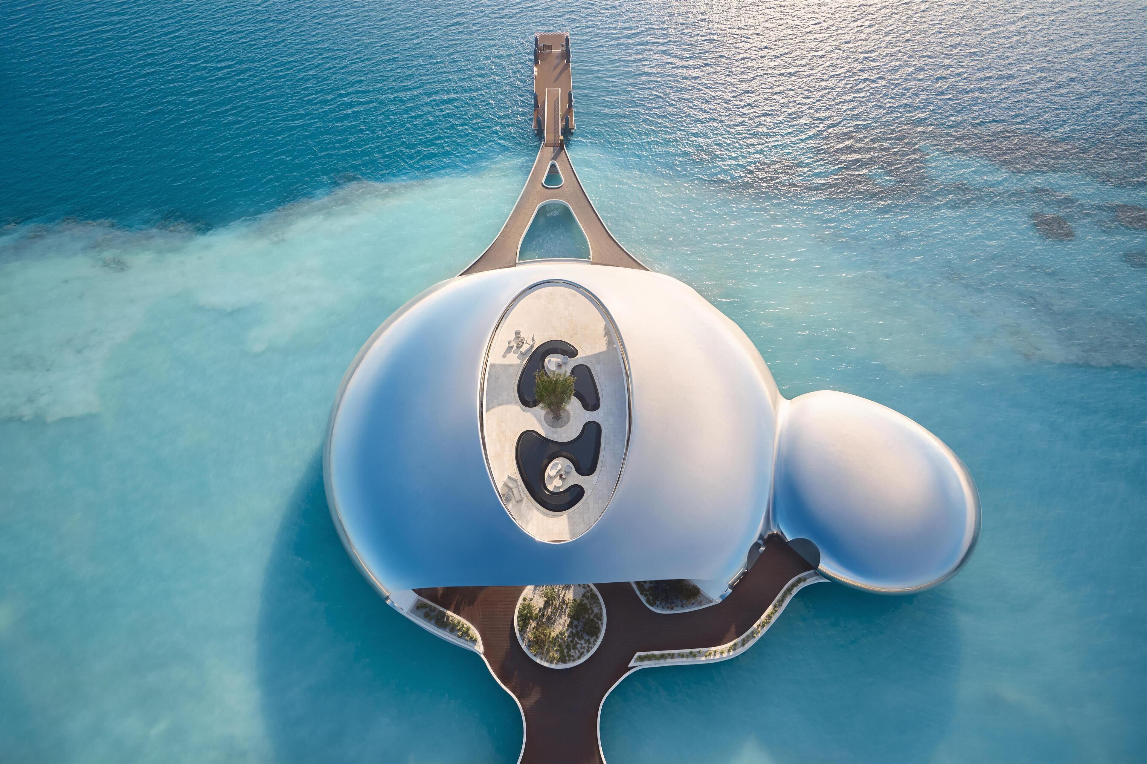 An aerial view of a unique, modern structure with a central circular feature, surrounded by a clear blue water body.