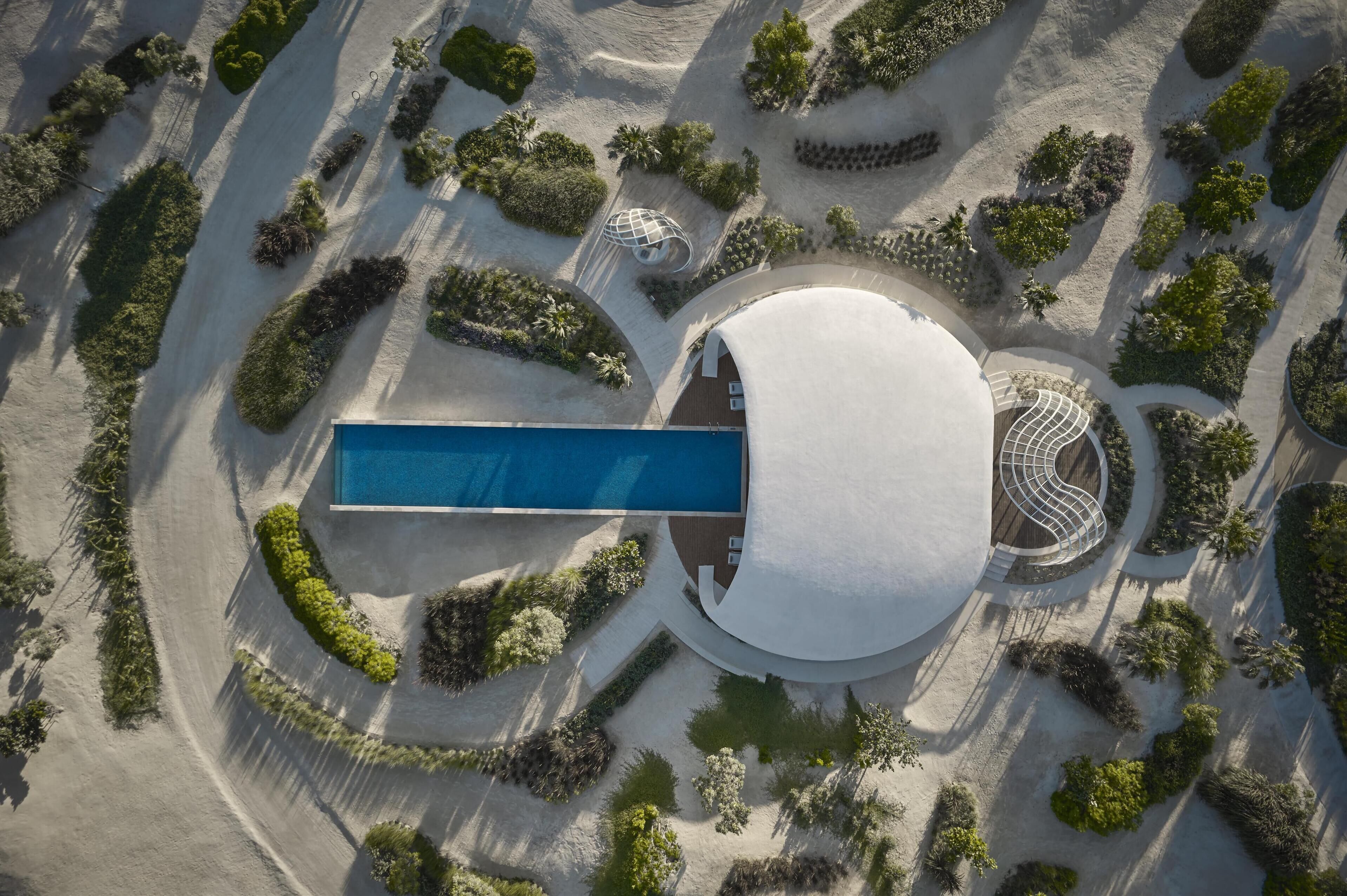 An aerial view of a modern building with a circular central area, surrounded by landscaped gardens and a long rectangular pool.
