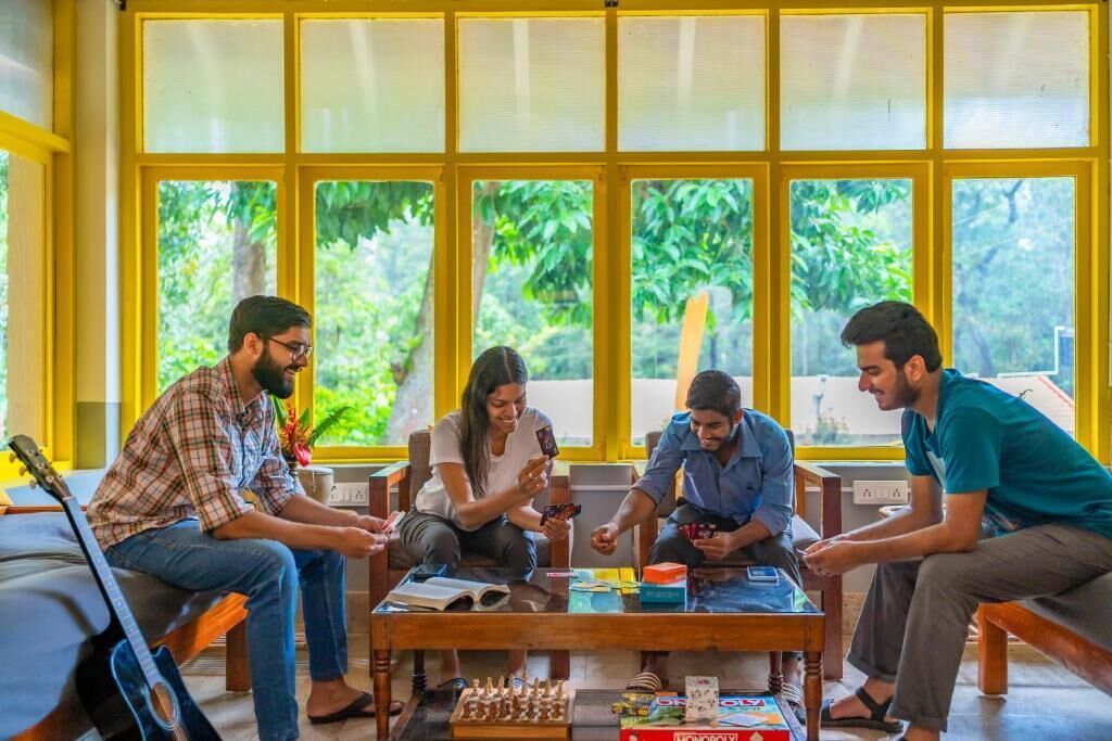 A group of people playing board games in a room with large windows and a guitar on a bench.