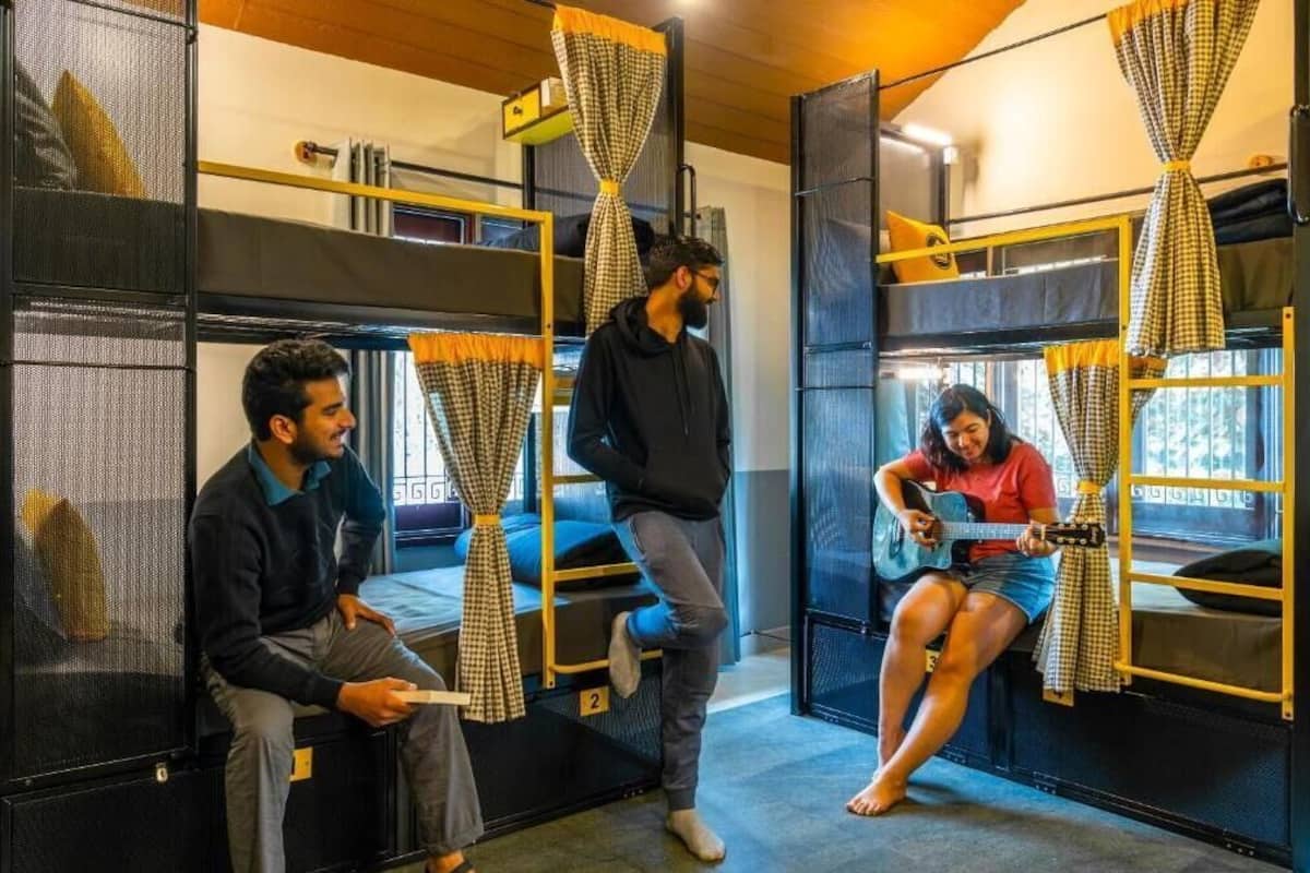 A hostel room with bunk beds, a window with curtains, and a person playing a guitar.