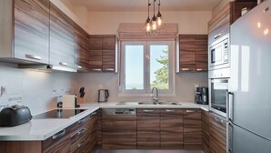 A modern kitchen with wooden cabinetry, stainless steel appliances, and a large window providing natural light. - Mili Serenity (Rethymno)