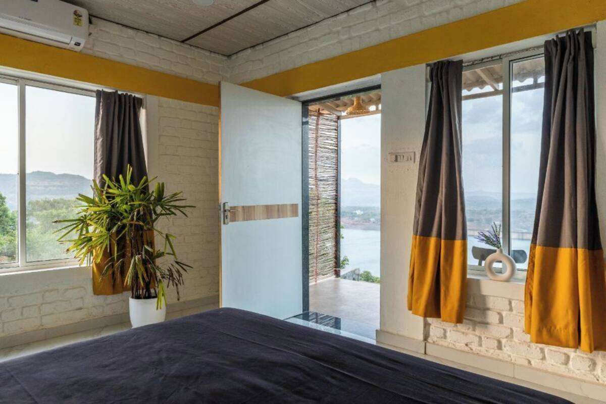 Un dormitorio con cama, una planta en maceta y vista a un cuerpo de agua a través de una ventana con cortinas.