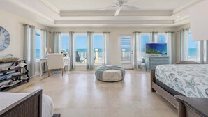 A spacious bedroom with a bed, a desk, a chair, a television, and a balcony with a view of the ocean. - High Tide Estate | Team Joseph Ellen (Ponte Vedra Beach)