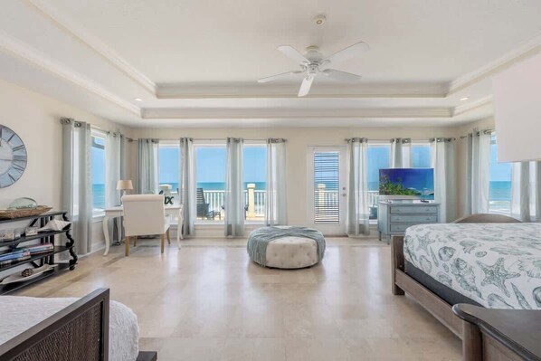 A spacious bedroom with a bed, a desk, a chair, a television, and a balcony with a view of the ocean.