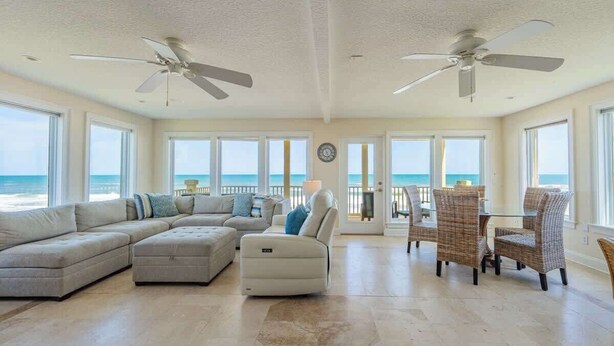 A spacious living room with a large sectional sofa, a chaise lounge, and a dining area with wicker chairs. There are ceiling fans and large windows offering a view of the ocean. - High Tide Estate | Team Joseph Ellen (Ponte Vedra Beach)