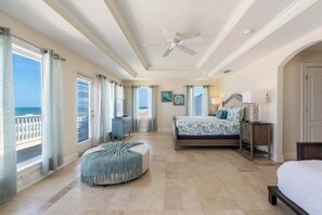 A bedroom with a bed, a nightstand, a lamp, a ceiling fan, and a view of the ocean through large windows. - High Tide Estate | Team Joseph Ellen (Ponte Vedra Beach)