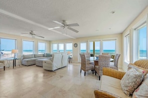 A spacious living area with a large window overlooking the ocean, a ceiling fan, and a dining table with wicker chairs.