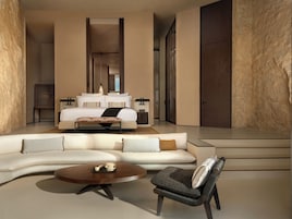 Una habitación de hotel moderna con un sofá grande, una mesa de centro y una silla. Hay una cama con una mesita de noche y una lámpara. La habitación tiene un ventanal y una puerta.