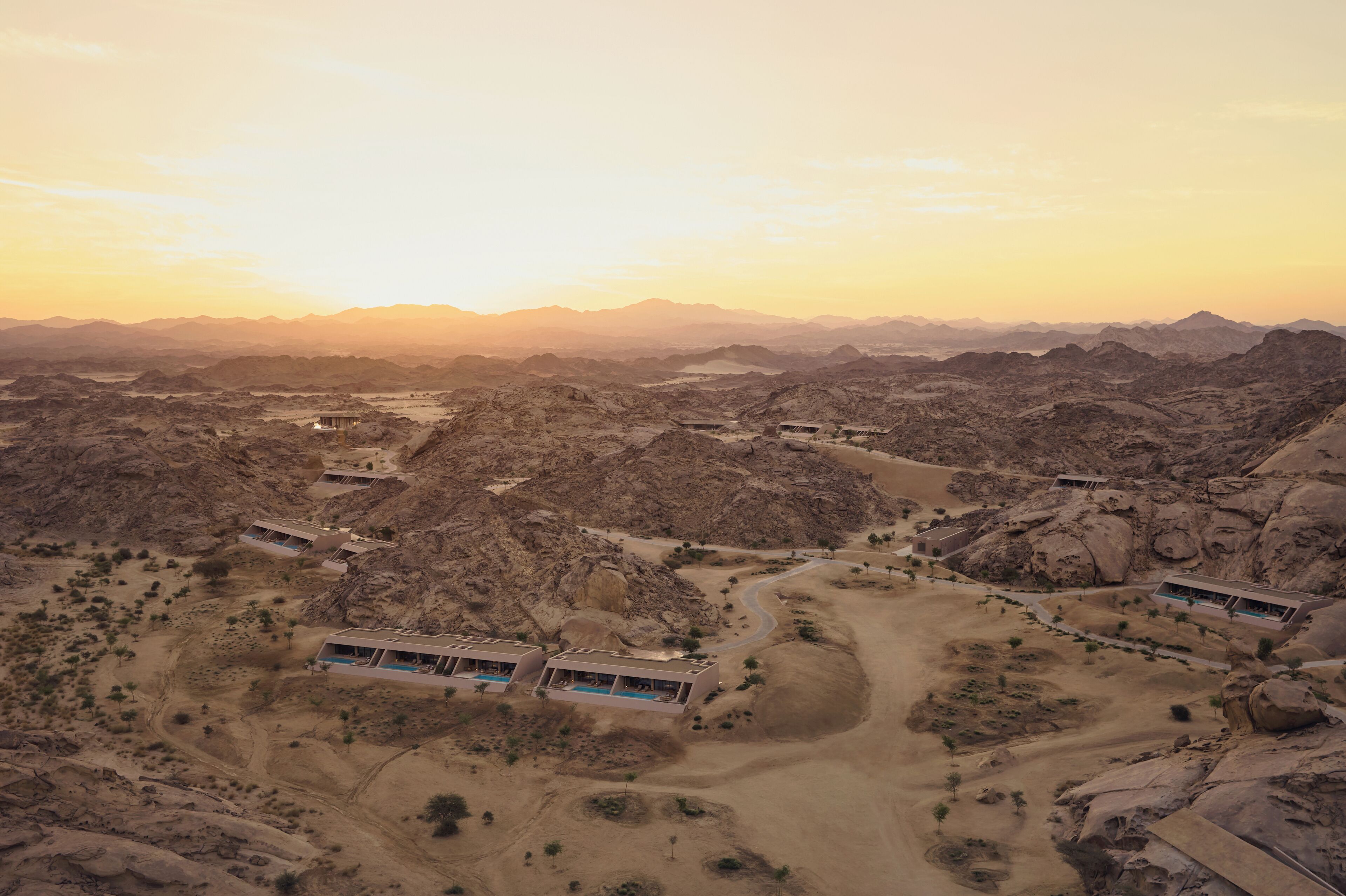 An aerial view of a desert resort with multiple buildings, including a large structure with a swimming pool, surrounded by rocky terrain and sparse vegetation.