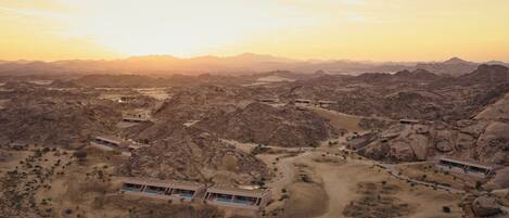 An aerial view of a desert resort with multiple buildings, including a large structure with a swimming pool, surrounded by rocky terrain and sparse vegetation.