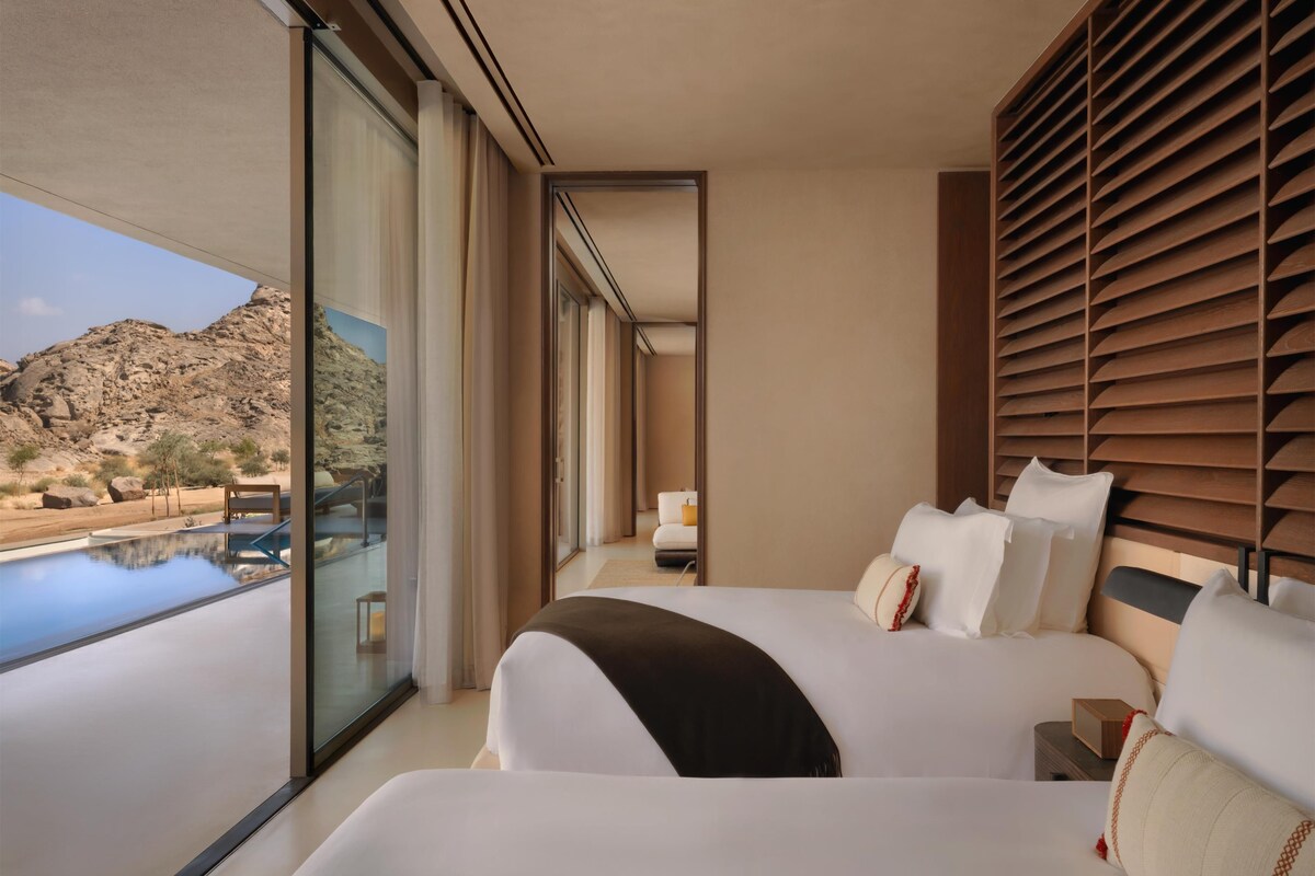 A modern hotel room with two beds, a large window, and a view of a rocky landscape.