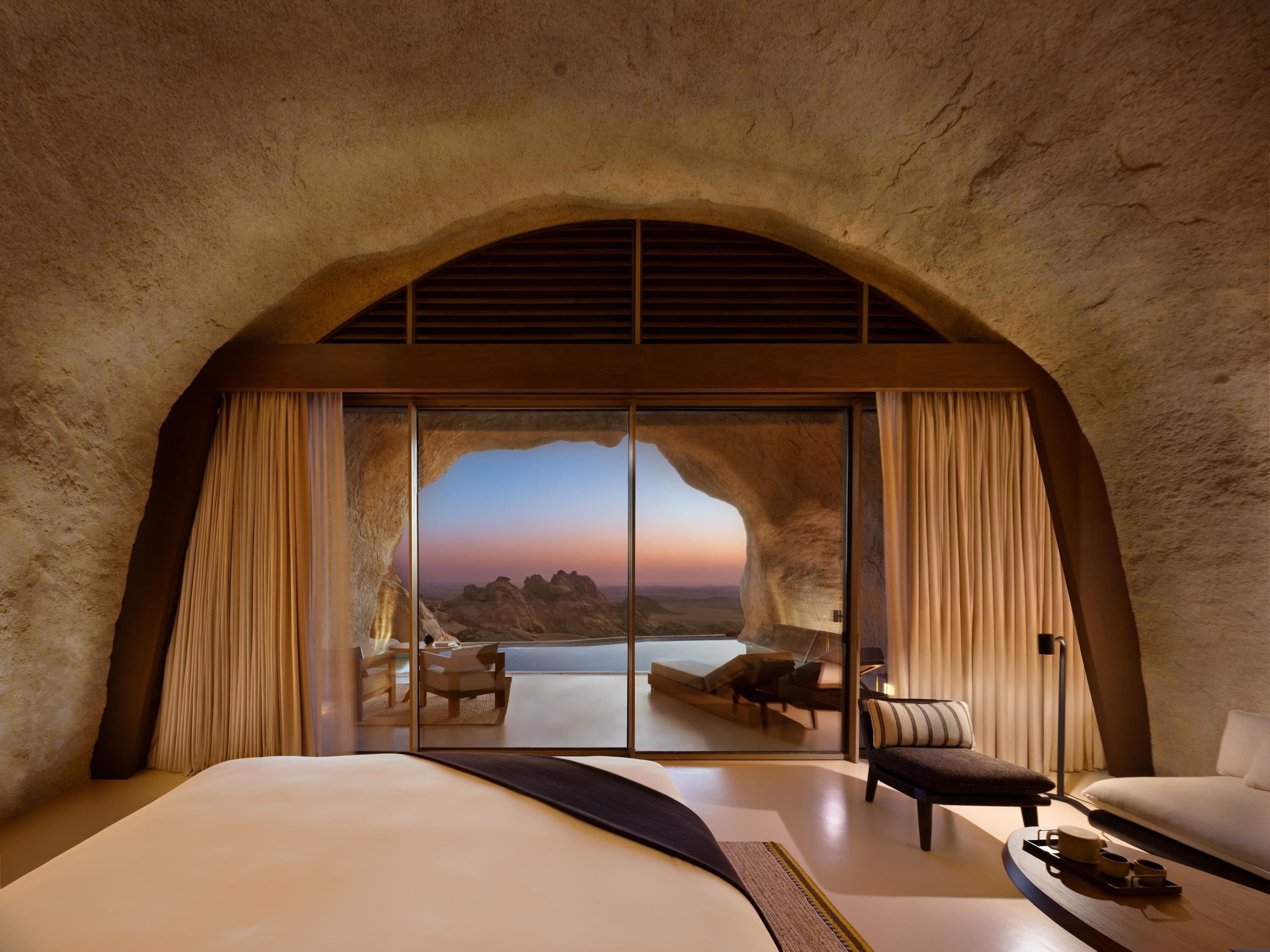 Un dormitorio con una cama grande, un área de descanso con una mesa, y vistas a un paisaje montañoso a través de un ventanal.