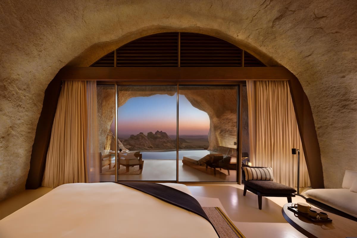 A bedroom with a large bed, a seating area with a table, and a view of a mountainous landscape through a glass door.