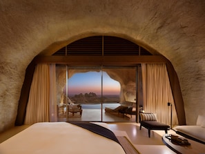 Ein Schlafzimmer mit einem großen Bett, einer Sitzecke mit Tisch und Blick auf eine gebirgige Landschaft durch eine Glastür.