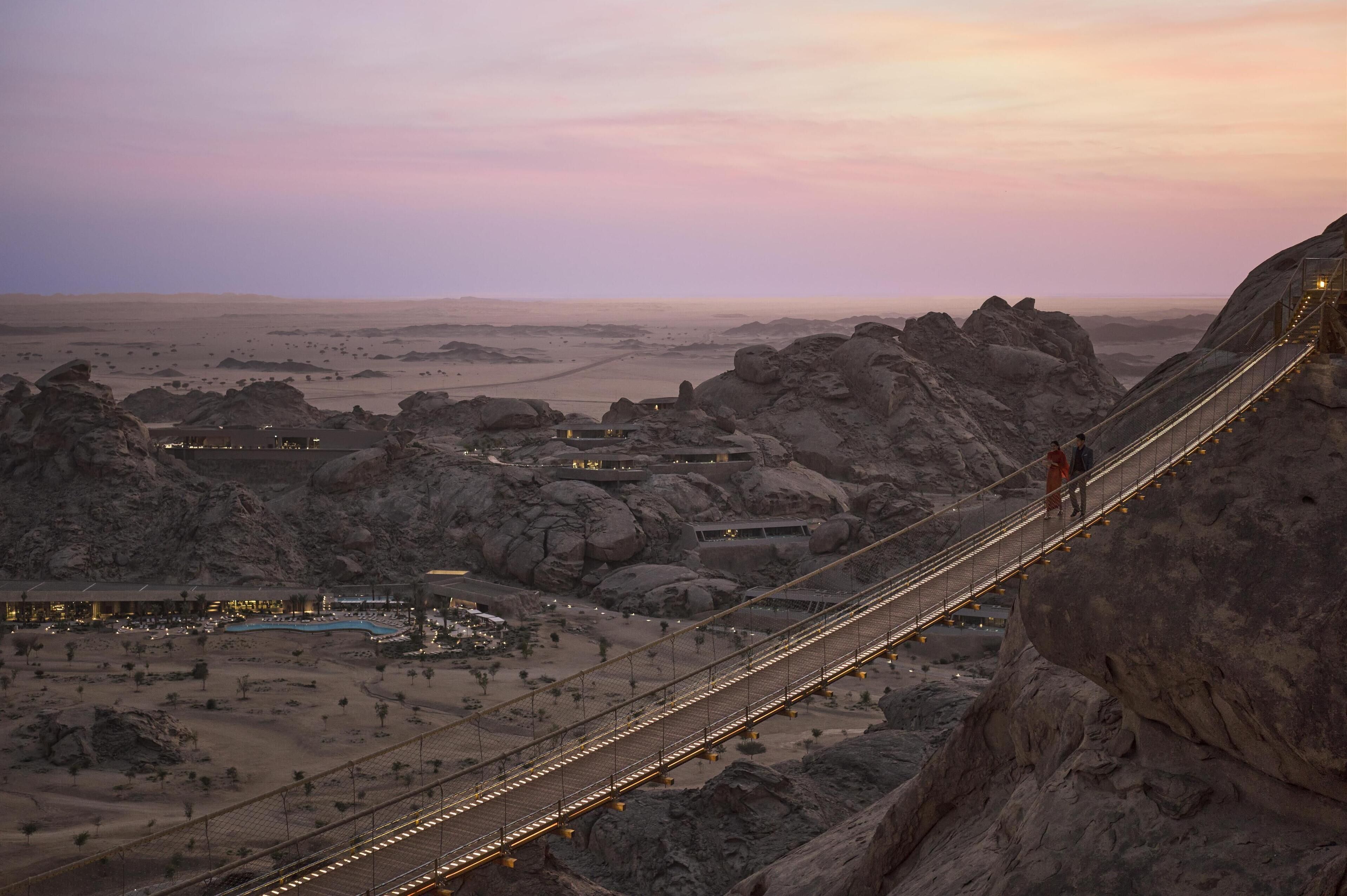 Un pont suspendu dans un paysage désertique, avec un terrain rocheux et des bâtiments situés au loin.