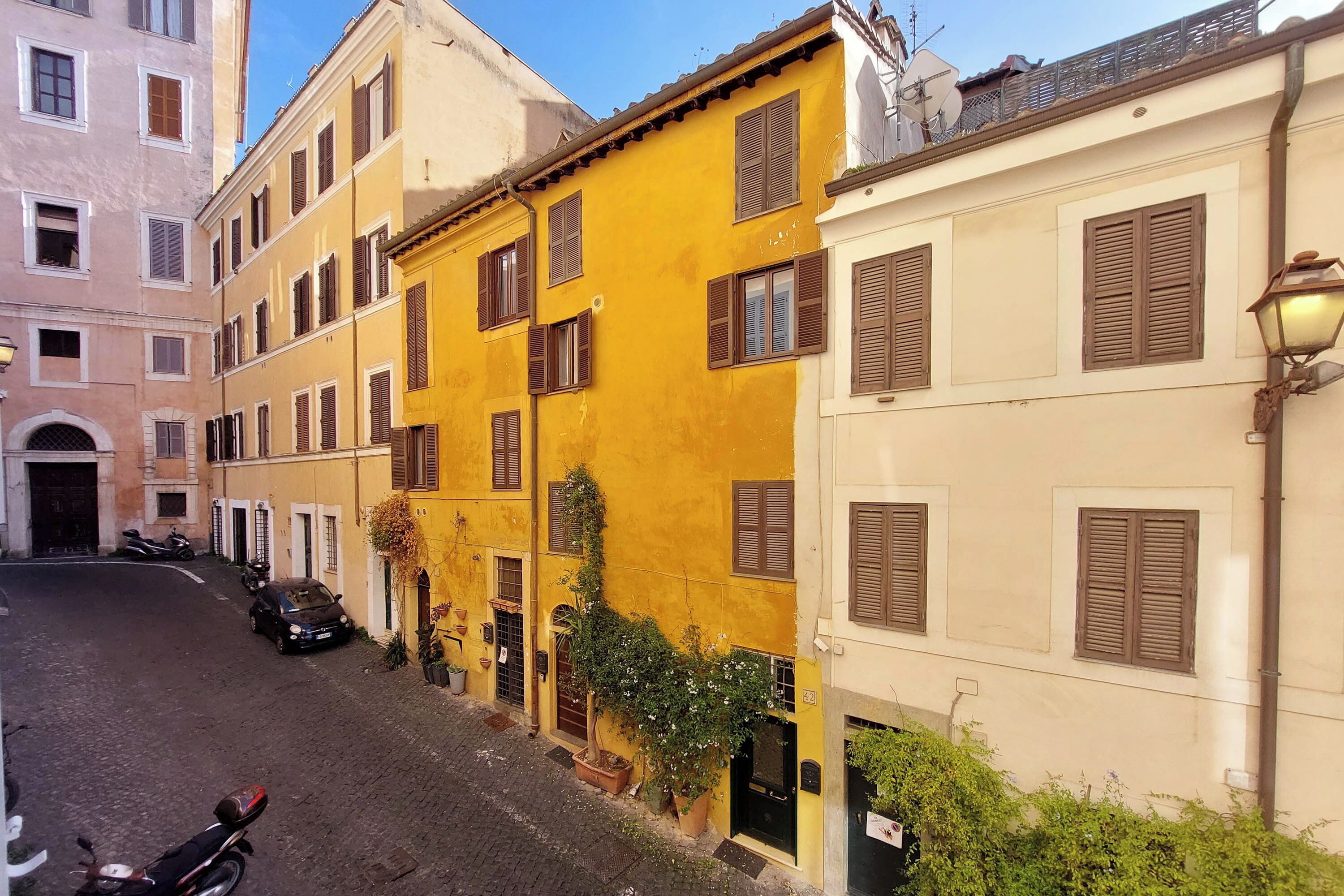 Una calle estrecha flanqueada por edificios, incluyendo un edificio amarillo con contraventanas y otro blanco también con contraventanas.