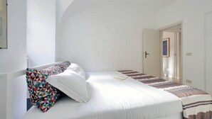 Una cama bien hecha con ropa blanca y almohadas estampadas, una pared blanca y una puerta que da a otra habitación.
