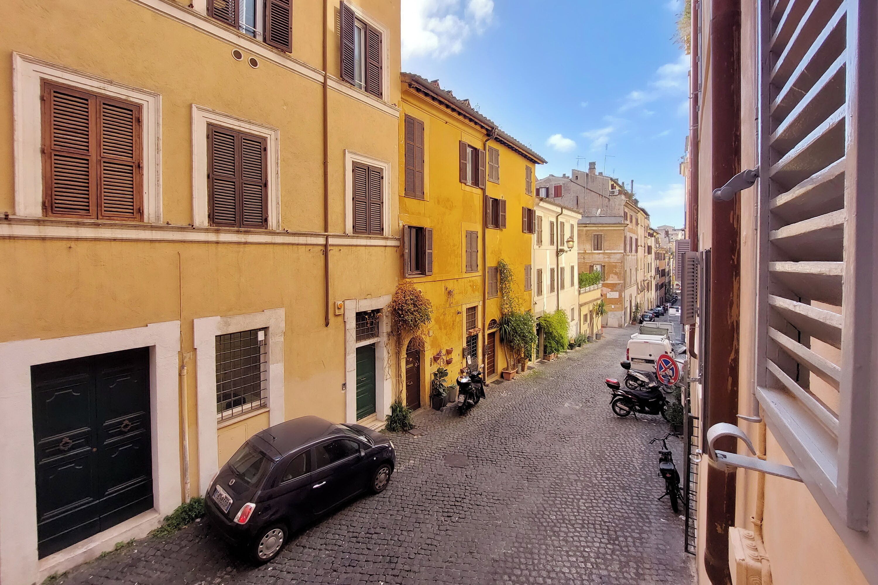 A narrow cobblestone street lined with yellow and white buildings, parked cars, and a motorcycle.