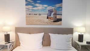 A hotel room with a bed, white pillows, a headboard, two bedside tables with lamps, and a beach-themed painting on the wall. - Fischer's Hus apartment 1<br>Beautiful apartment with terrace and access to the sea time (Büsum)