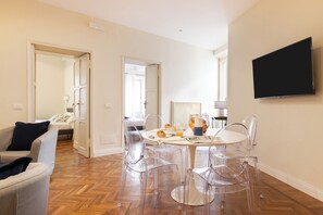 A modern living room with a wooden herringbone floor, a round dining table with clear chairs, a flat-screen TV mounted on the wall, and a sofa with a dark cushion. - Coppelle Charme (Rome)