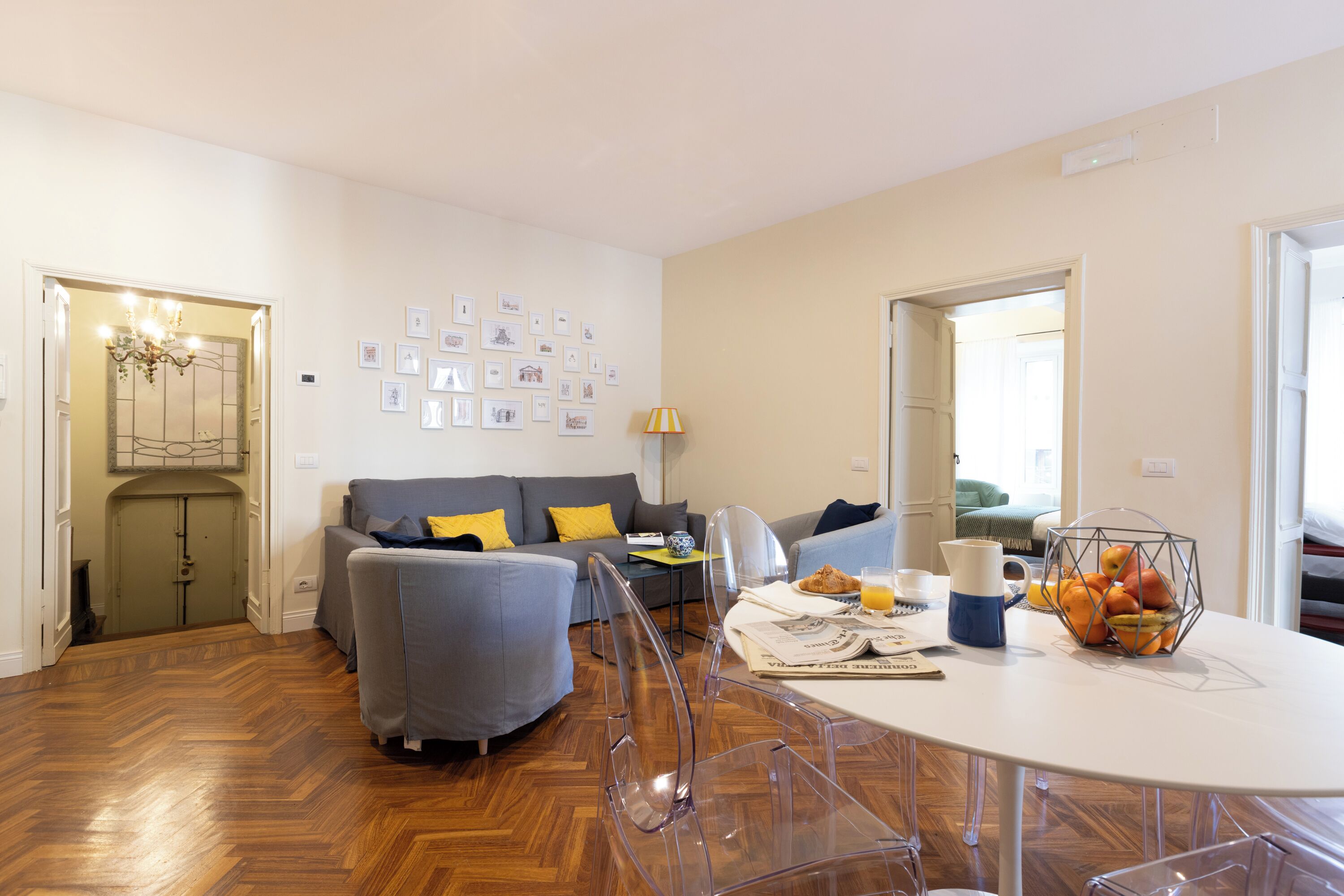 A modern living room with a grey sofa, a dining table with chairs, and a wooden herringbone floor.
