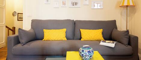 A living room with a grey sofa, yellow cushions, a vase, and framed pictures on the wall.