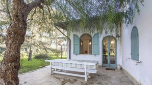 A rustic outdoor seating area with a bench and table, surrounded by a white building with green shutters and arched windows. - Villa Cardinale  in Salento with pinewood and swimming pool. Puglia. (Puglia)