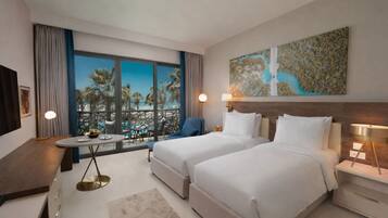 Ein Hotelzimmer mit zwei Betten, einem Balkon mit Blick auf den Pool, einem Esstisch und einem Gemälde an der Wand.