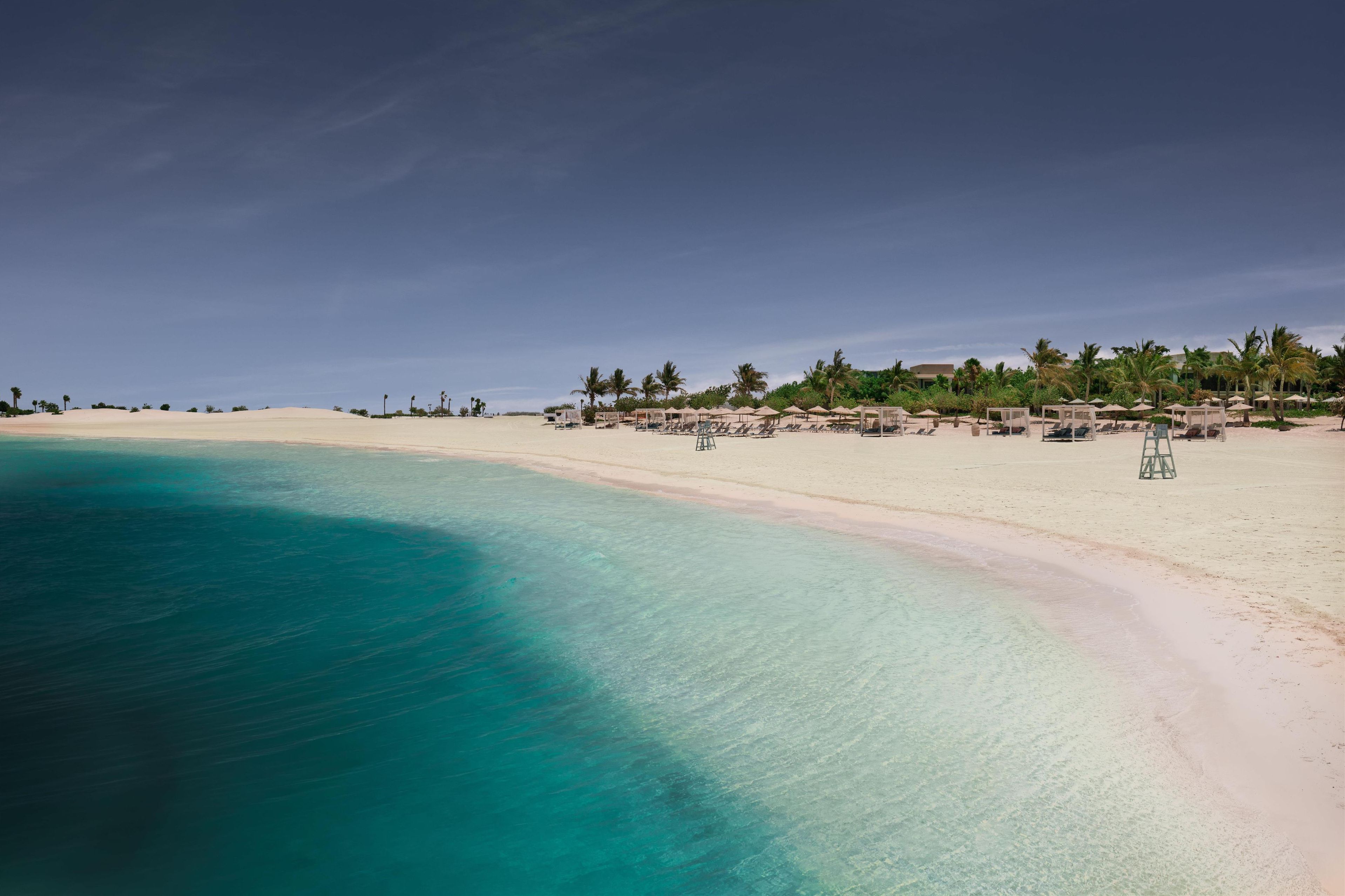 A beach with clear turquoise water, white sand, and palm trees. There are sun loungers and umbrellas along the shore.