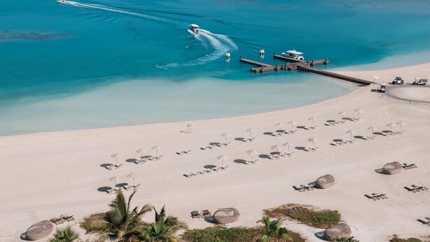 Eine Vogelperspektive zeigt einen Strand mit klarem, blauem Wasser, ein Jetski sowie eine Pier mit Liegestühlen und Sonnenschirmen.