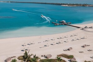 Oversigtsbillede af en strand med klart blåt vand, en jetski og en moler med liggestole og parasoller.