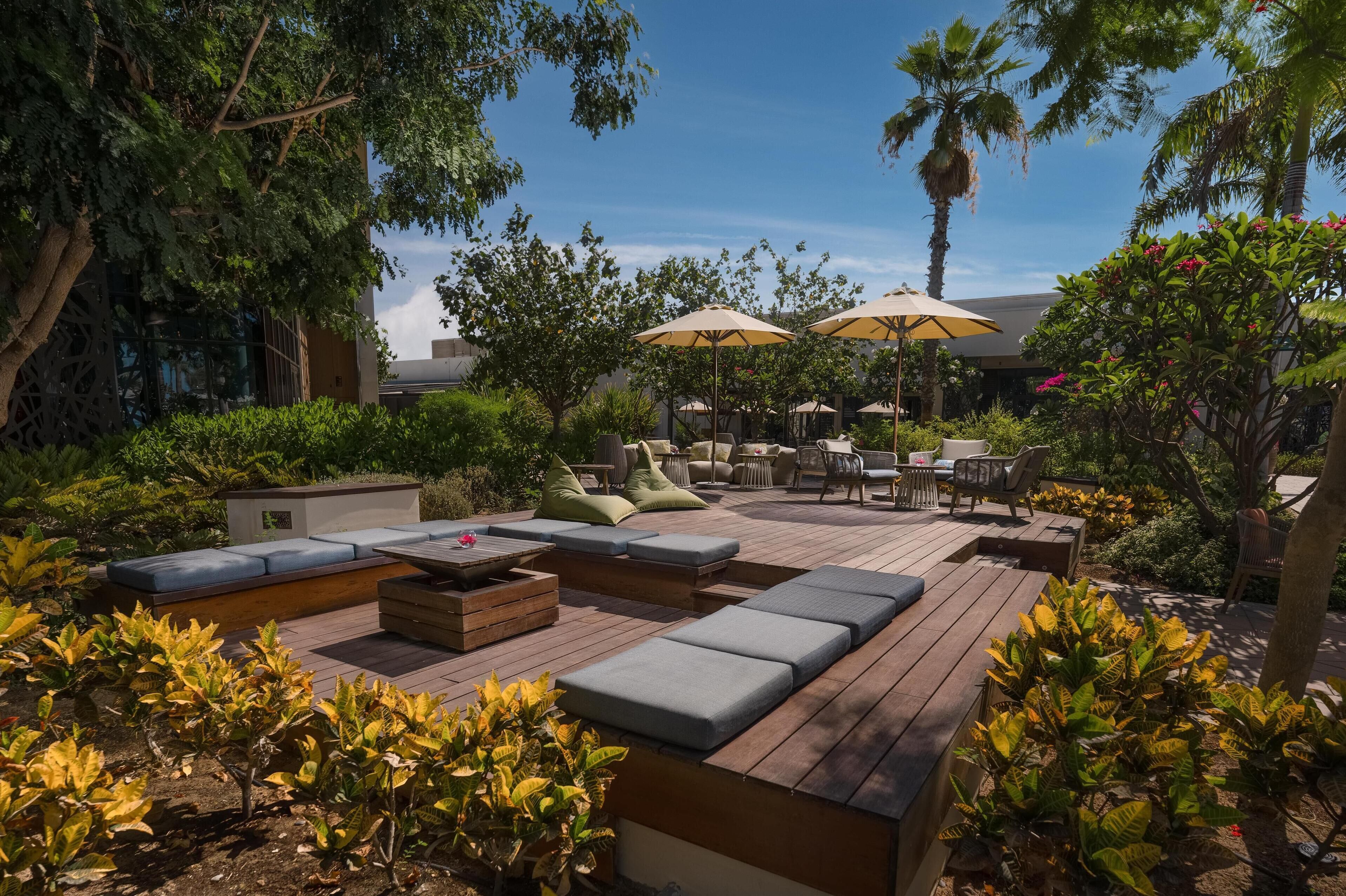 A wooden deck with outdoor seating, including sofas and chairs, surrounded by lush greenery and palm trees.